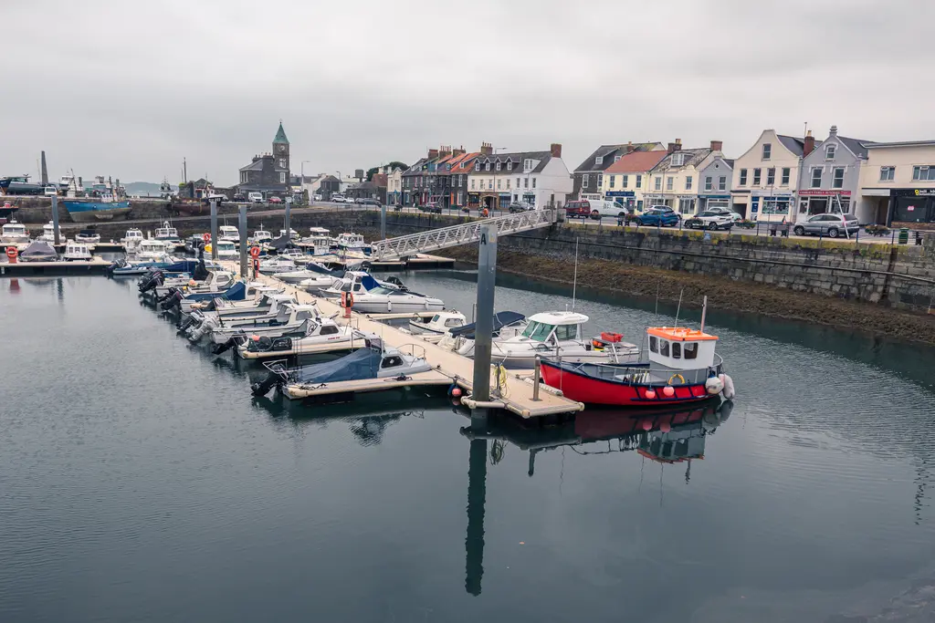 A marina with various boats docked, surrounded by charming buildings and a calm harbor on a cloudy day.