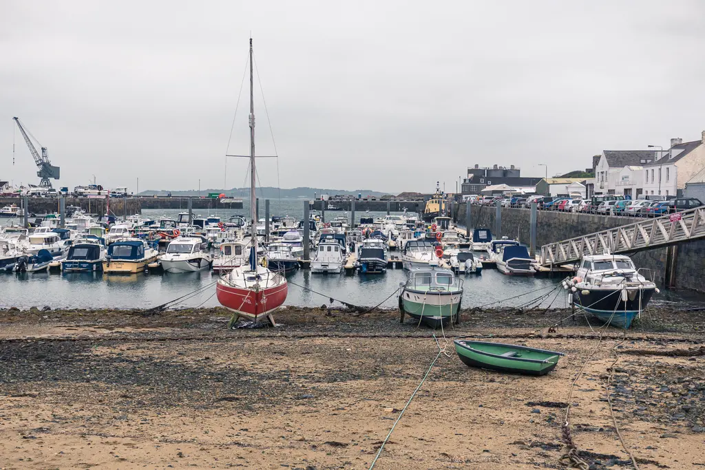 A coastal harbor with numerous boats moored, sandy shore in the foreground, and a cloudy sky above.