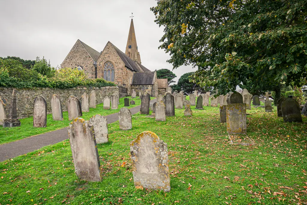A historic church surrounded by a graveyard, featuring tombstones and lush green grass under a cloudy sky.