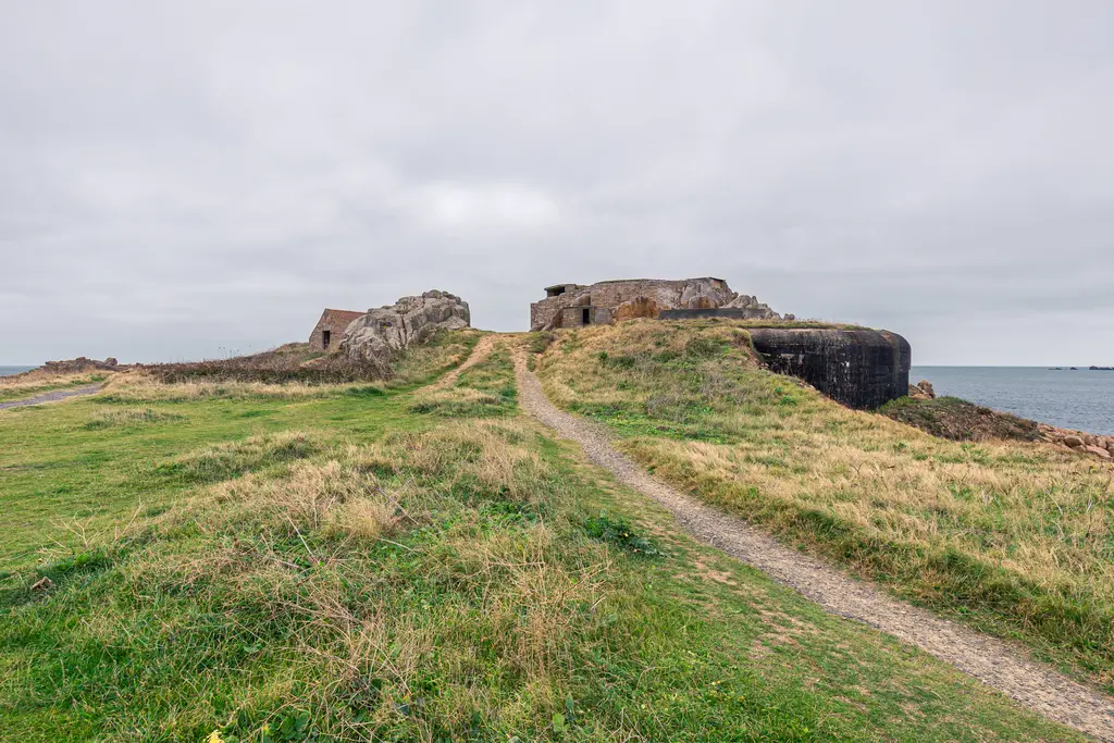 A historic coastal fortification on a grassy landscape with a winding path leading to stone structures and the sea beyond.