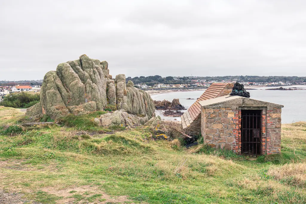 A rugged rock formation beside a weathered stone building overlooking the sea, with a distant coastal town in the background.