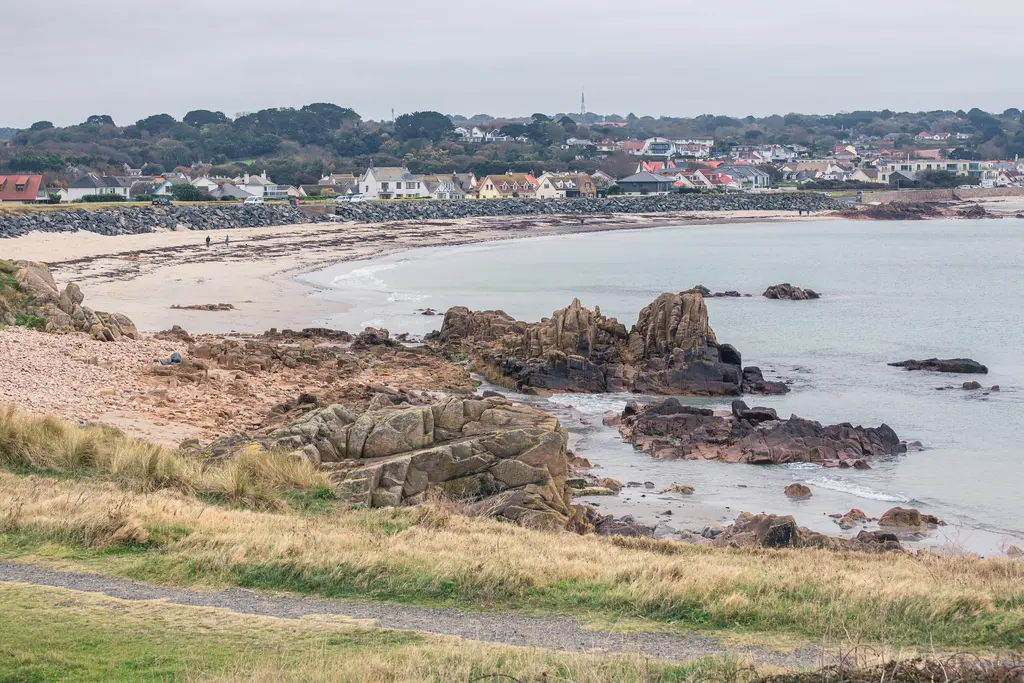 A coastal view featuring rocky formations, sandy beach, and a quaint village backdrop under overcast skies.