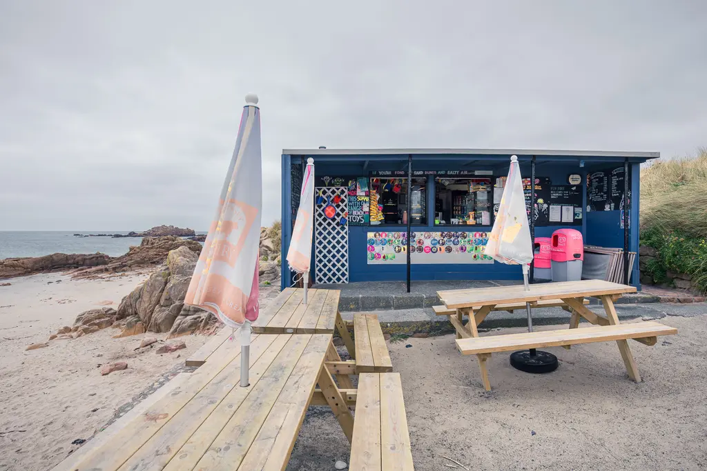 A beachside kiosk with picnic tables, colorful decorations, and flags, set against rocky seascape and overcast sky.