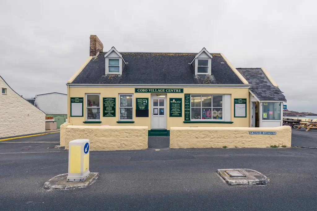 A cream-colored village center building with a slate roof, featuring shop windows and green signage in a coastal town setting.