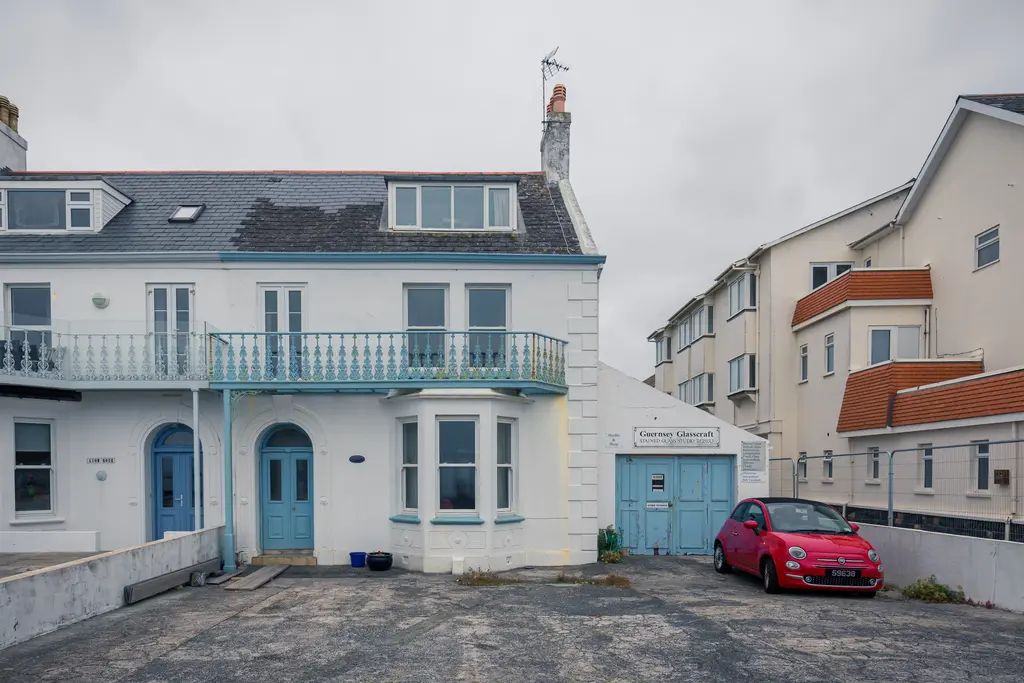 A charming coastal house featuring blue accents, surrounded by modern buildings and a red car parked nearby.