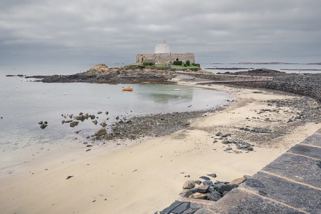 A coastal view featuring a sandy beach, rocky shore, and a historical fort on the horizon under a cloudy sky.