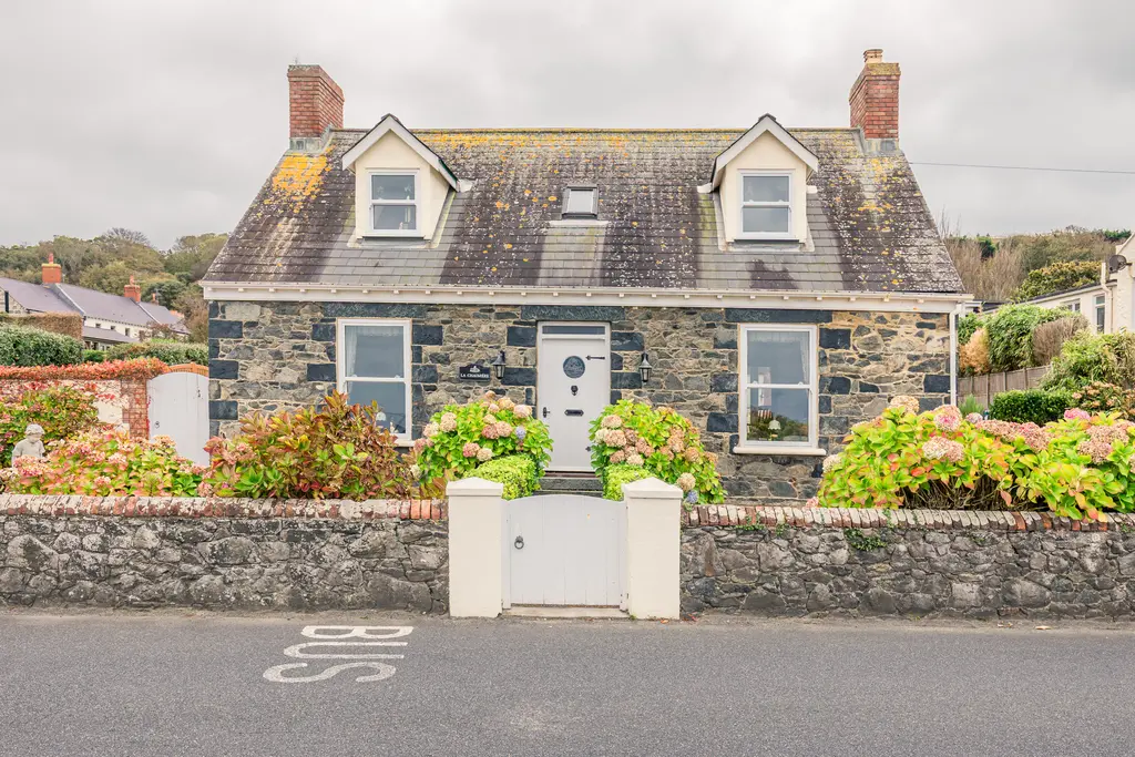 A charming stone cottage with a slate roof, surrounded by colorful flowers and a low stone wall. A serene street scene.