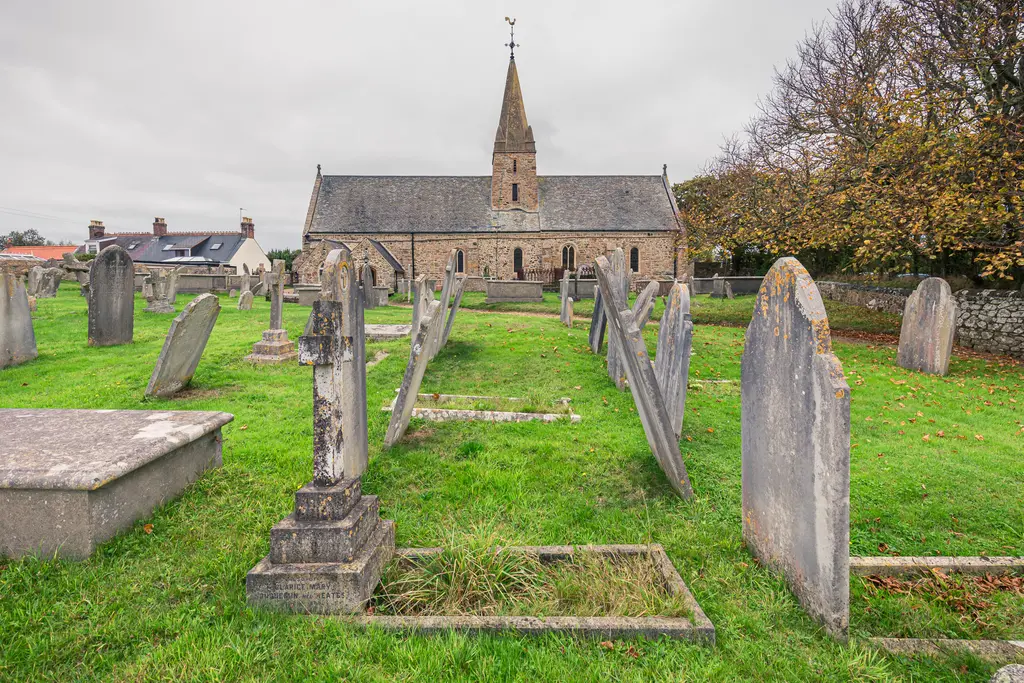 A graveyard with weathered tombstones in front of an old stone church, surrounded by grass and trees on a cloudy day.