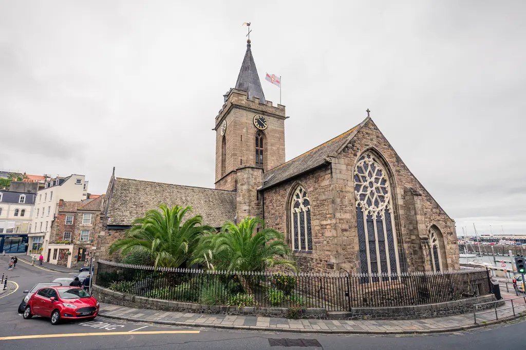 An historic stone church with a tall clock tower, surrounded by palm trees and urban buildings, in a coastal setting with overcast skies.
