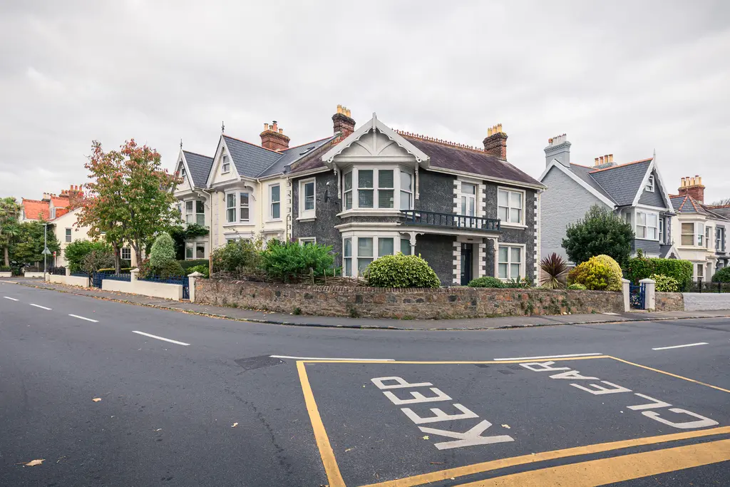 A charming residential street featuring Victorian-style houses surrounded by greenery, with a cloudy sky above.
