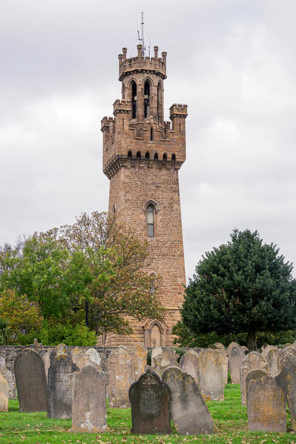 A historic stone tower surrounded by gravestones and lush greenery under a cloudy sky, evoking a serene atmosphere.