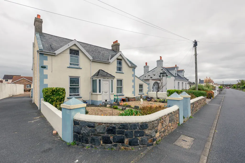A charming house with a pale yellow exterior and gray roof, surrounded by a stone wall and garden, alongside a quiet street.