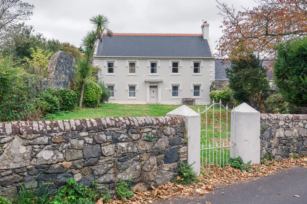 A charming white house surrounded by greenery and a stone wall, featuring a gated entrance and autumn foliage.