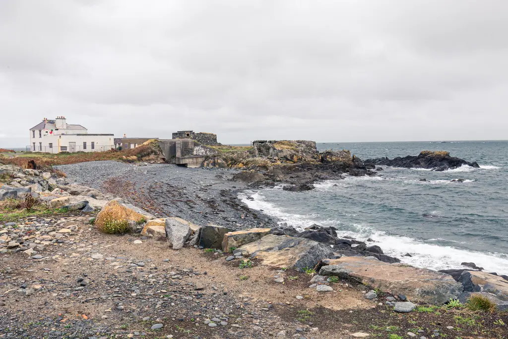 A rocky coastal scene featuring a white building, pebbled shoreline, and waves crashing against rocks under a cloudy sky.