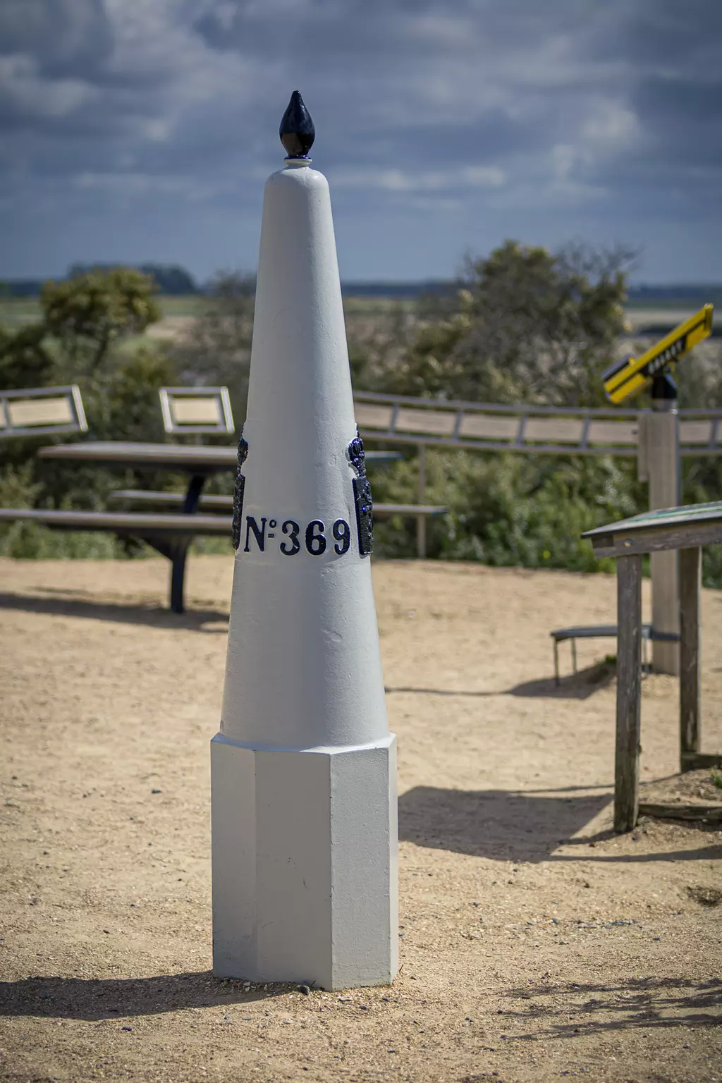 Ein konischer Steinpfeiler mit der Aufschrift N°369, umgeben von sandigem Boden und Bänken im Hintergrund.