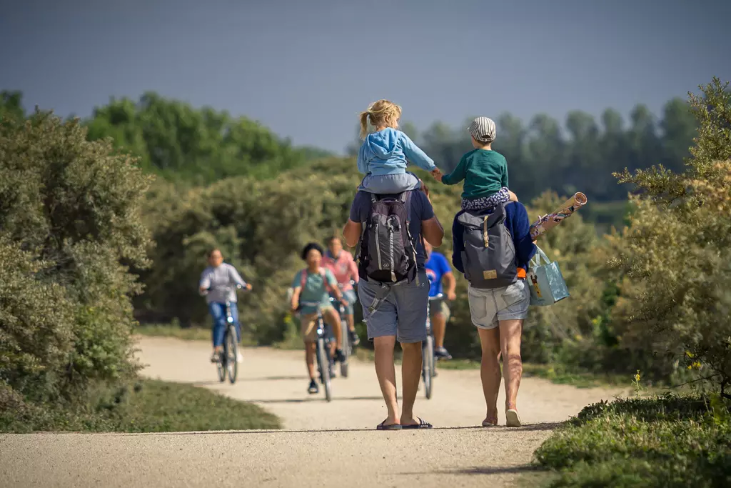 Familie geht auf einem Weg; ein Erwachsener trägt ein Kind auf den Schultern, ein weiteres Kind sitzt auf einem anderen Rücken.