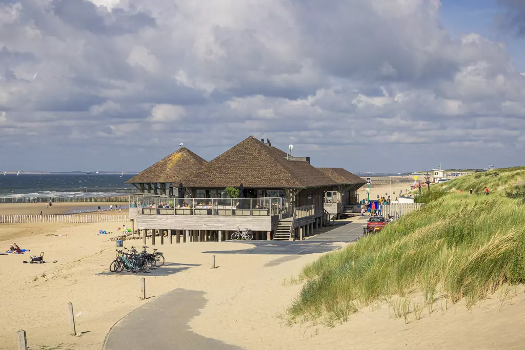 Ein Holzgebäude mit einem steilen Dach steht am Strand, umgeben von Sanddünen. Fahrräder sind geparkt und Menschen entspannen.