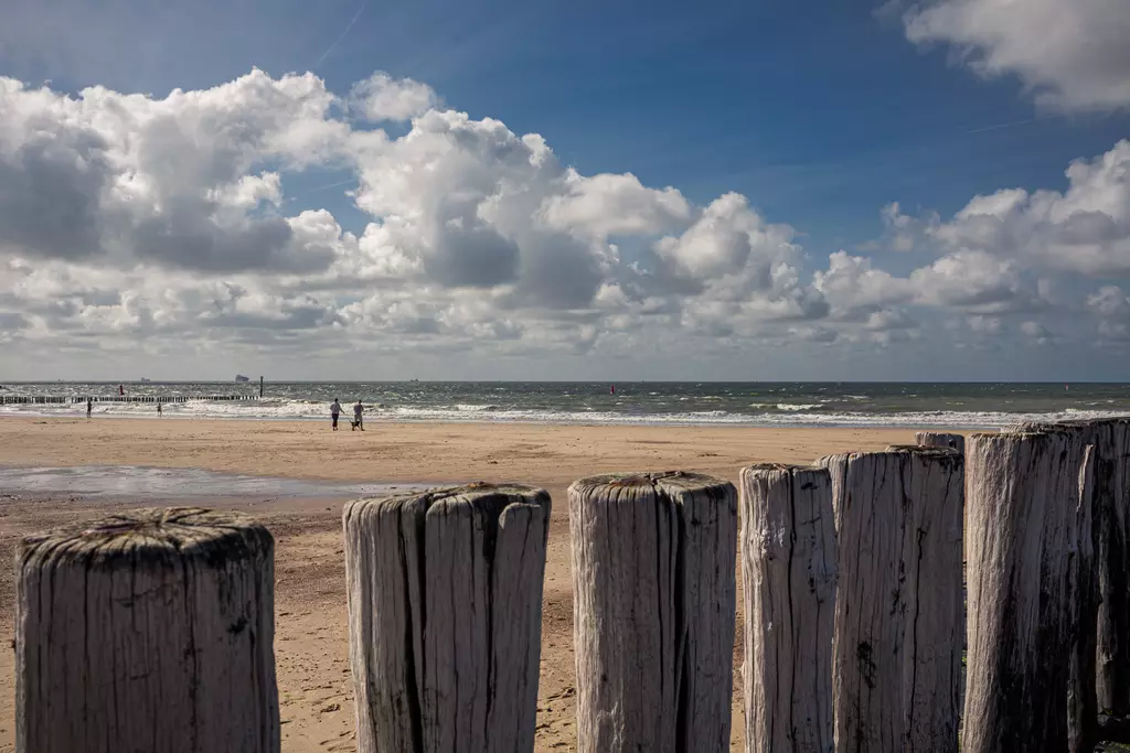 Holzpfähle stehen im Vordergrund, während Strandbesucher am Meer entlang gehen. Über dem Wasser schieben sich Wolken am blauen Himmel.