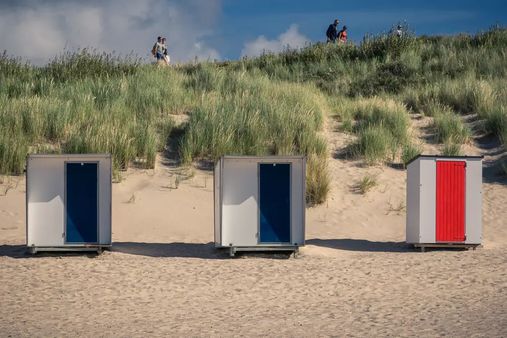 Drei Strandkabinen stehen auf einem sandigen Strand. Im Hintergrund sind Menschen zu sehen, die auf einem Hügel spazieren.