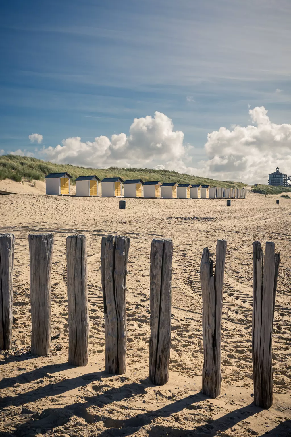 Holzpfähle stehen im Sand, im Hintergrund sind mehrere bunte Strandhäuser und sanfte Dünen unter einem strahlend blauen Himmel zu sehen.