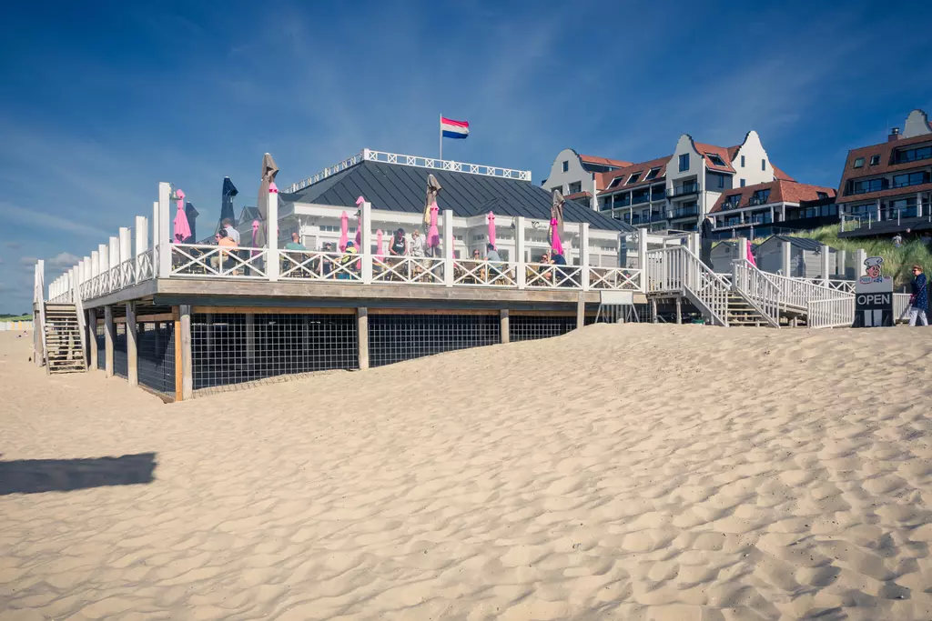 Ein Strandrestaurant aus Holz mit pinken Sonnenschirmen, umgeben von Sanddünen und großen Gebäuden im Hintergrund.