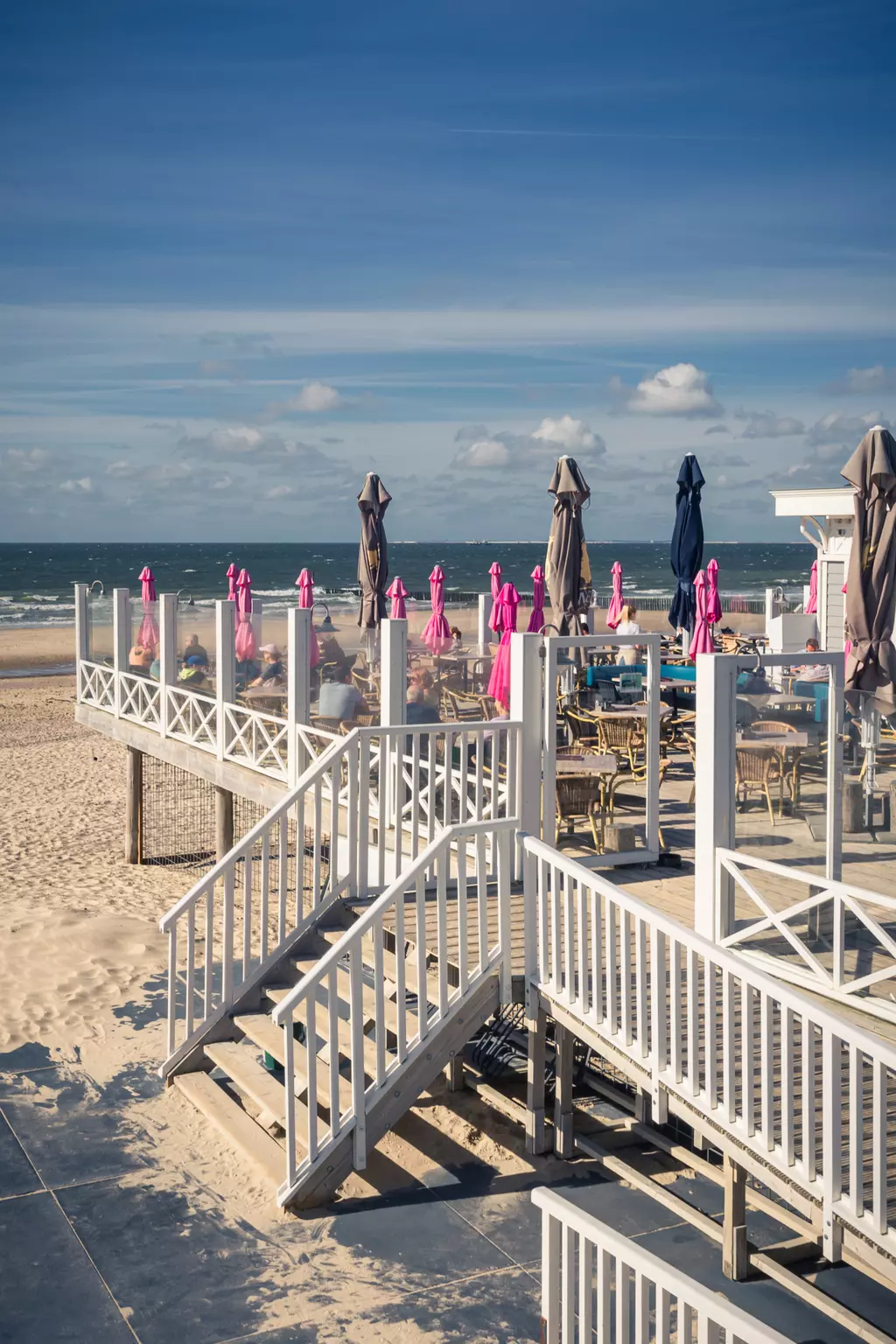 Eine Holzterrasse mit weißen Geländern und rosa Sonnenschirmen, Blick auf das Meer unter einem blauen Himmel mit wenigen Wolken.