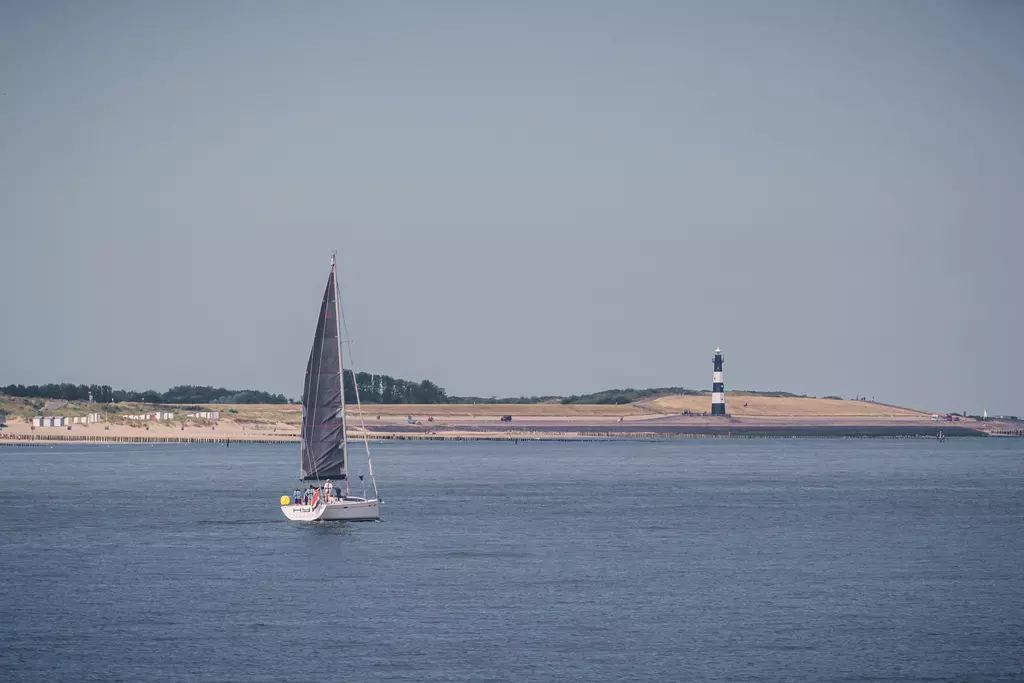 A sailboat with a black sail glides across the water, with a lighthouse in the background along the coastline.