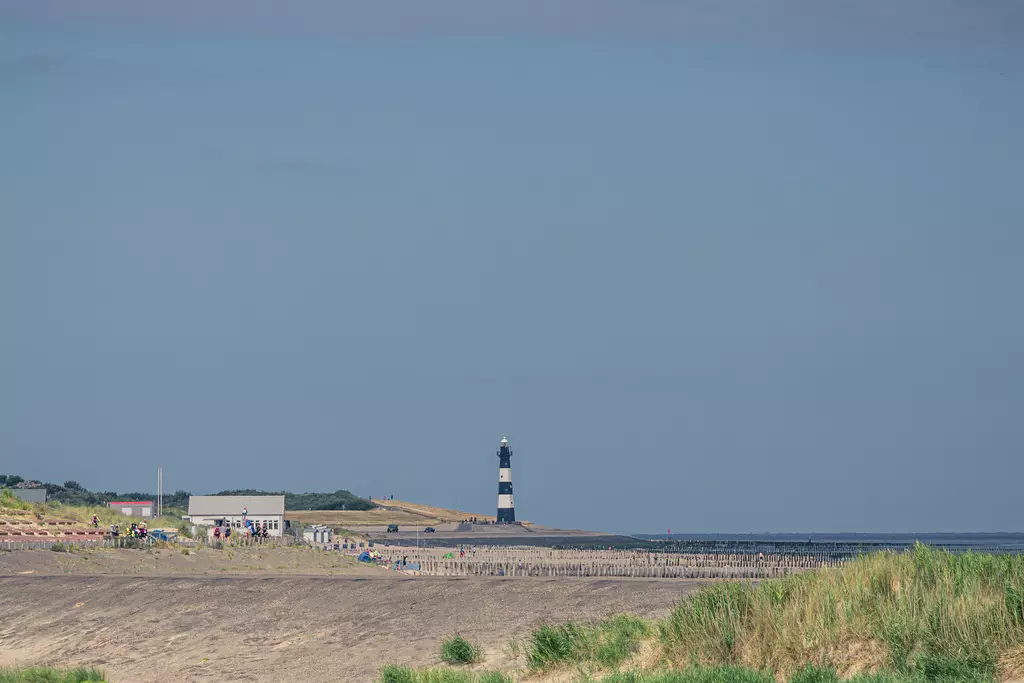 A beach with many people, a cluster of buildings, and a black and white lighthouse in the background.