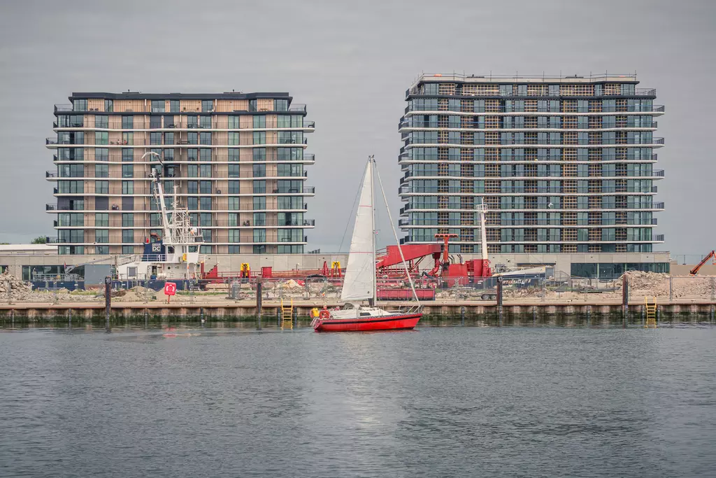 A small sailboat with a red hull passes by modern high-rise buildings along the harbor.