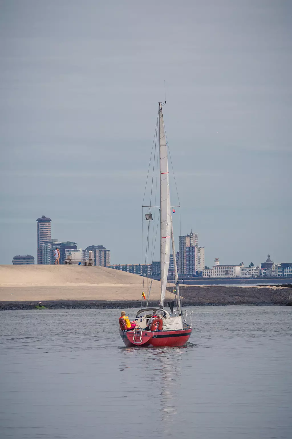 A sailboat with two people glides on calm water towards a coastal city featuring modern buildings in the background.