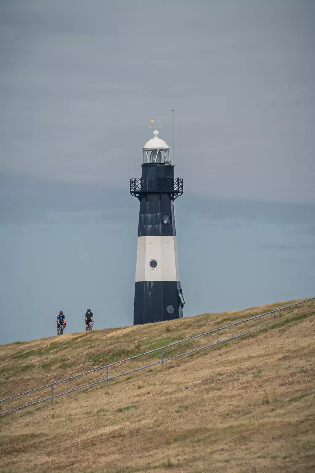 Two cyclists stand on a hill with a black and white lighthouse in the background under a cloudy sky.