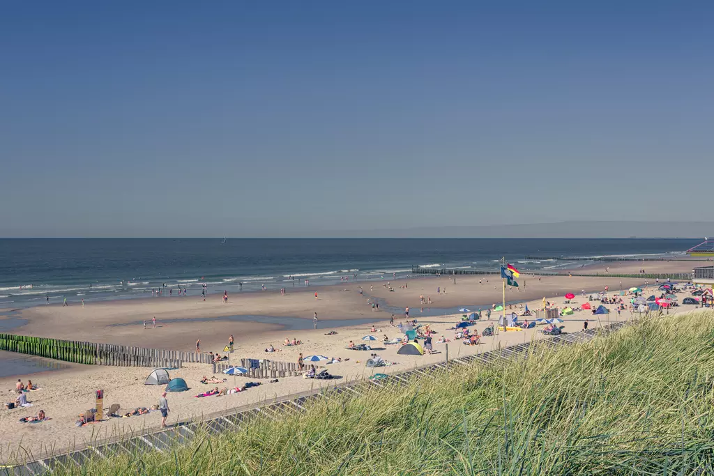 A wide beach with sunbathers, deck chairs, and colorful umbrellas, surrounded by gentle dunes.