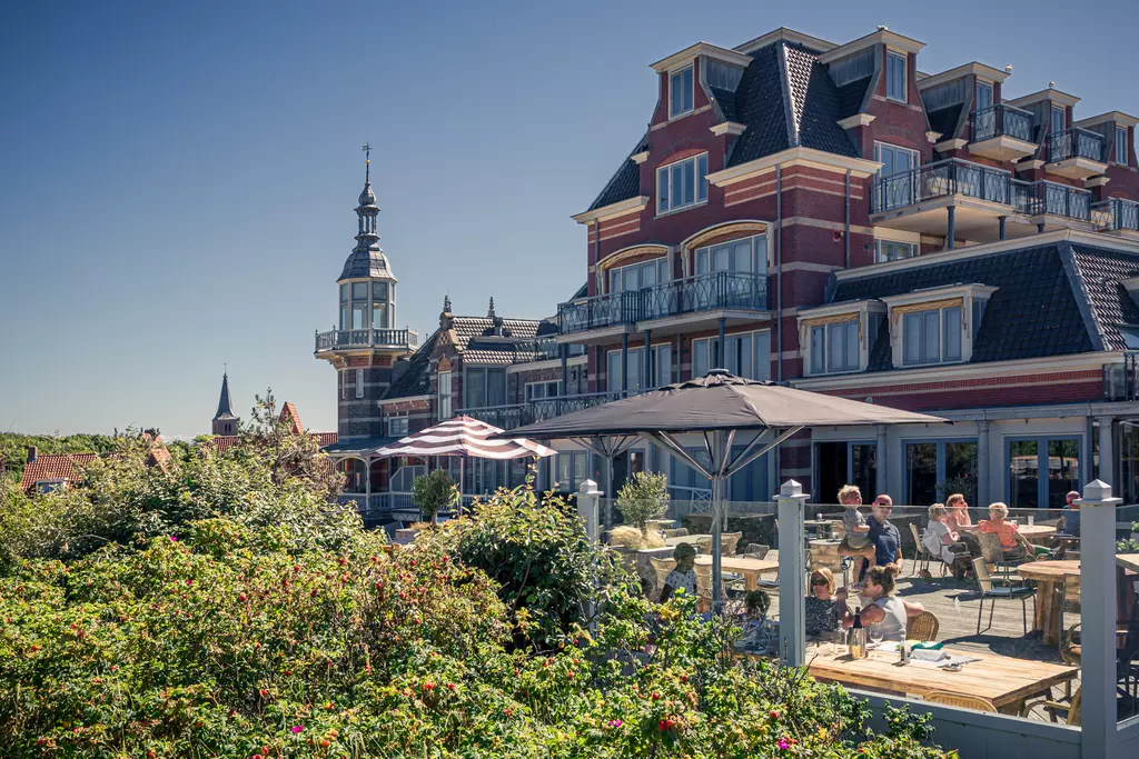 Blooming plants surround a terrace in front of a stylish building with a tower, where guests relax in sunny weather.