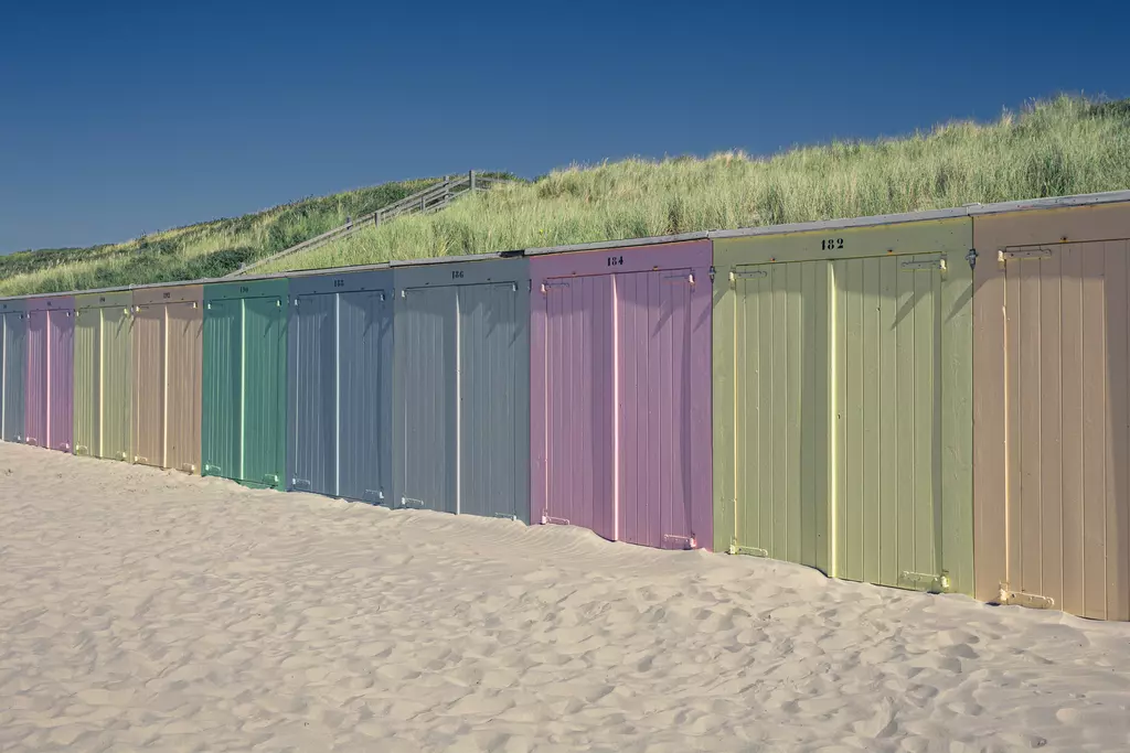 Colorful beach huts lined up on bright sand, surrounded by gentle green dunes in the background.