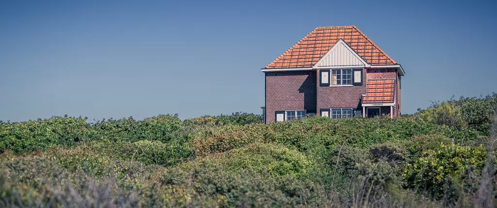 A red house with a pointed roof stands among green bushes and shrubs against a clear sky.