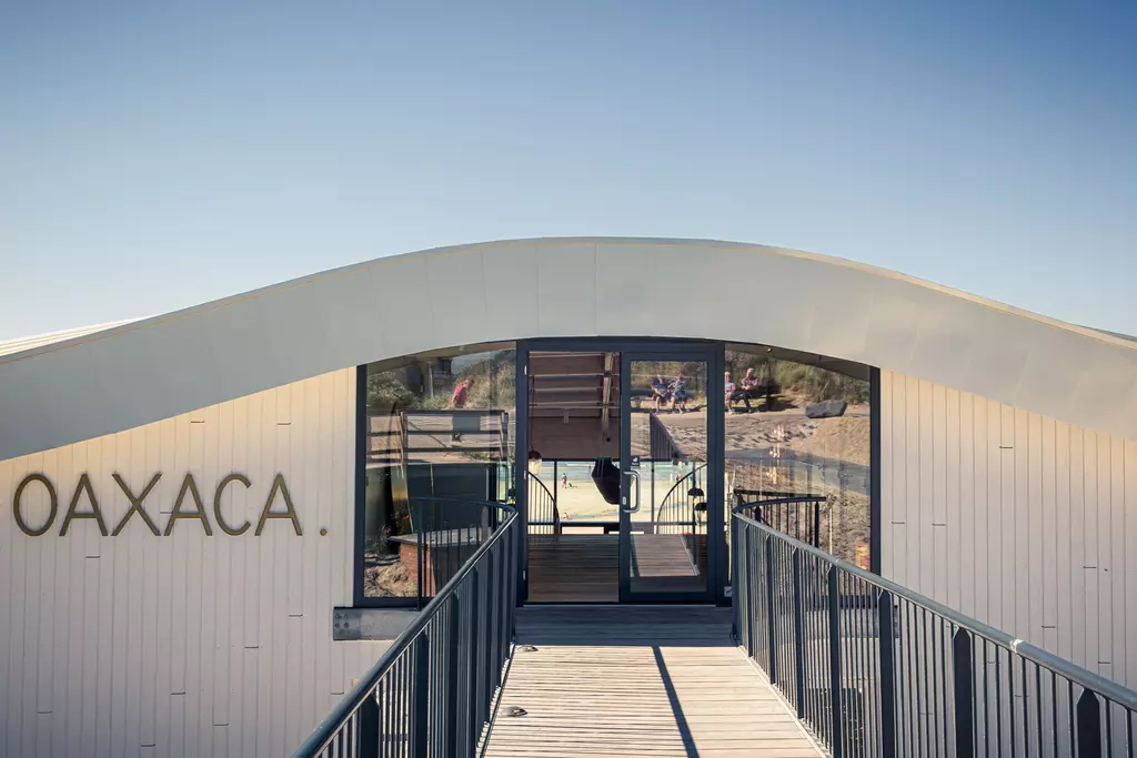 A modern building with a curved roof labeled Oaxaca, featuring a wooden walkway leading to the entrance.