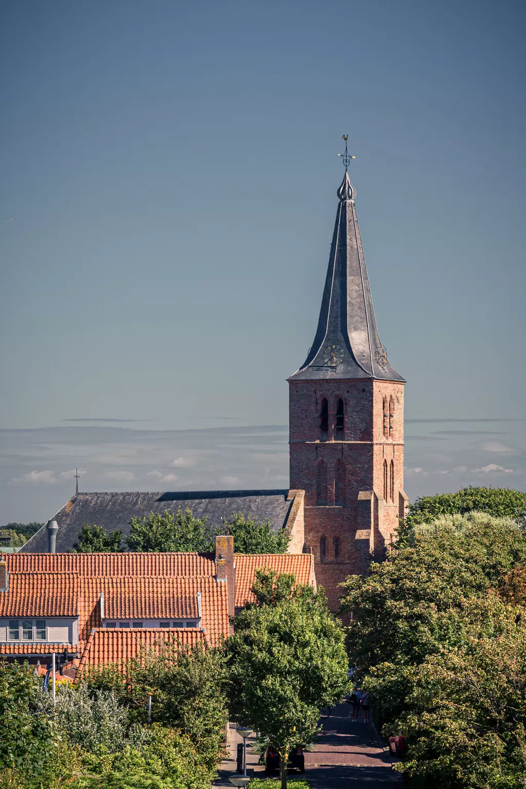 A church with a distinctive pointed tower rises above buildings and trees under a clear sky.