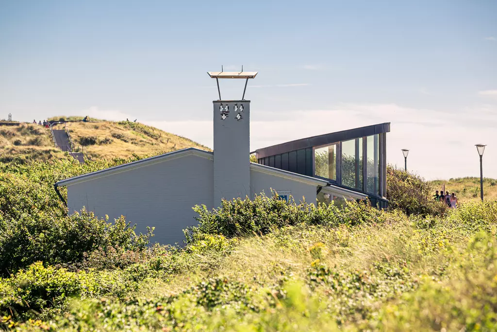 A modern building with a glass facade is nestled among hilly dunes and greenery. People walk along a path in the background.