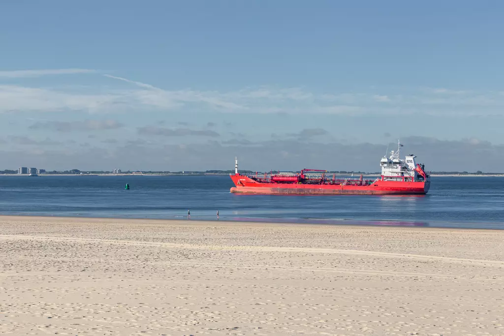 A red ship sails on calm water, with a sandy beach in the foreground and a lightly clouded sky in the background.