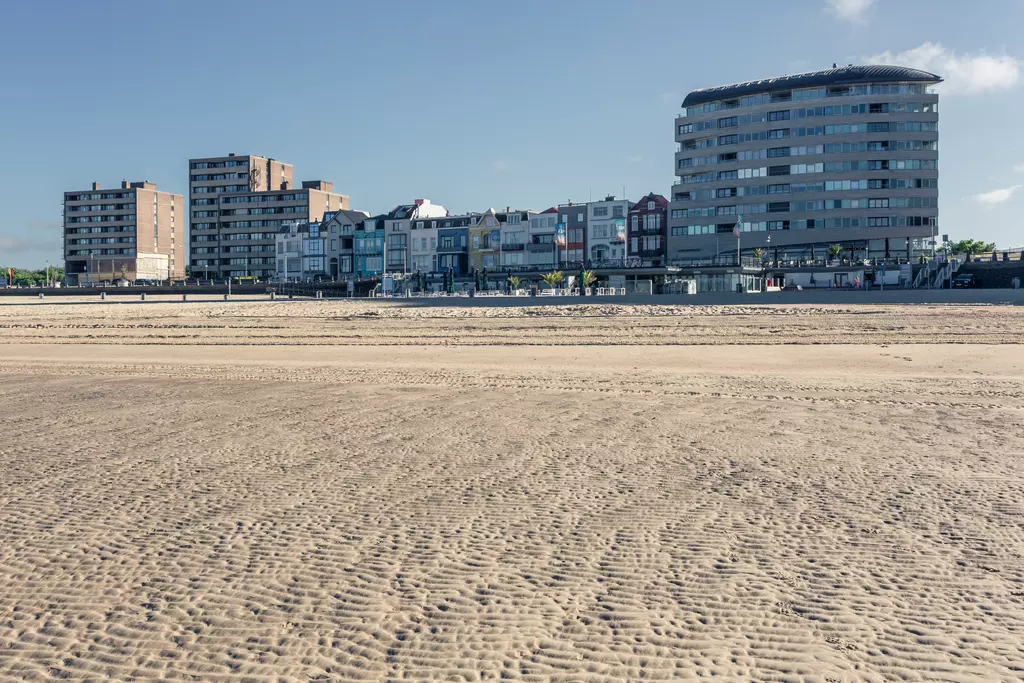 Sandy beach with patterned tracks, backed by multi-story buildings along the coast. Clear sky, calm atmosphere.