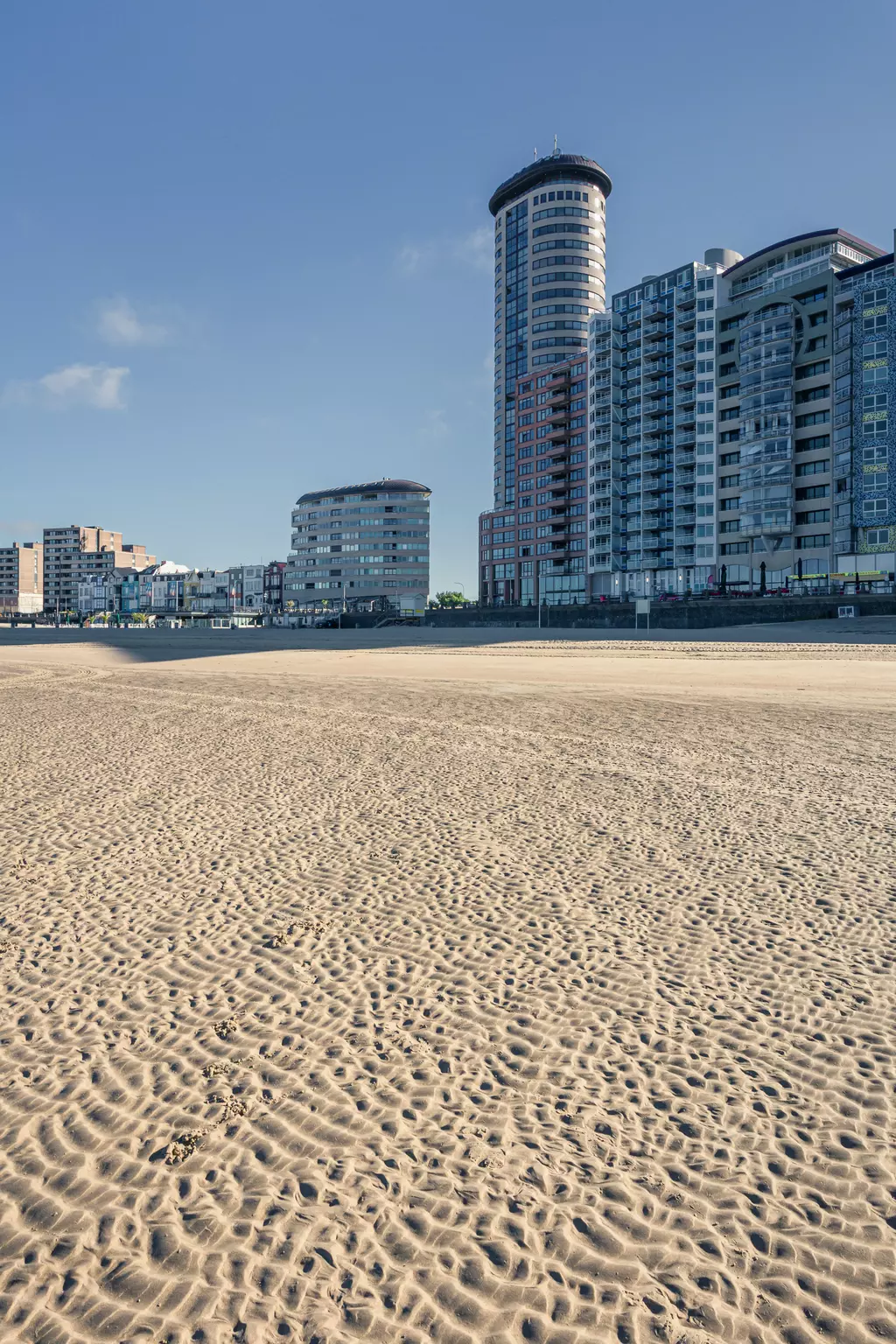 Sandy beach with fine patterns in the foreground, high-rise buildings in the background, blue sky with a few clouds.