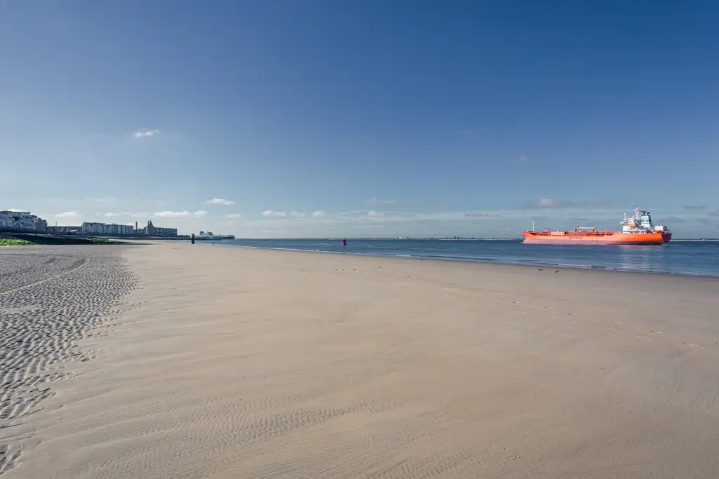 A serene beach with fine sand, featuring calm waters and an orange cargo ship in the distance under a clear blue sky.