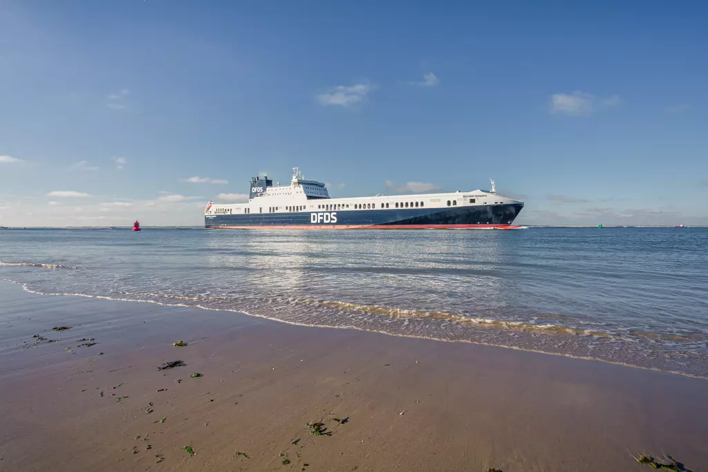 A ferry is passing on calm waters, with a clear blue sky and sandy beach in the foreground.