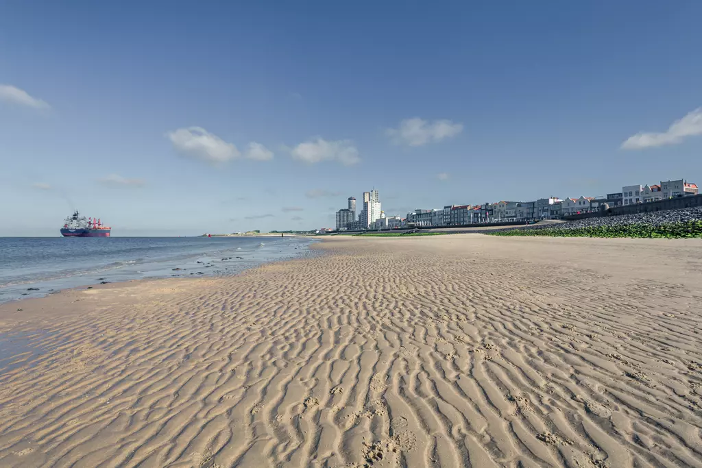 Sandy beach with wave patterns, a ship in the water, and a cityscape featuring modern buildings in the background.