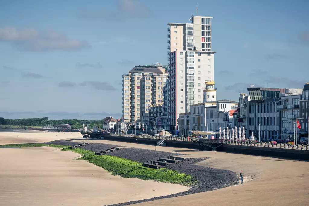 A beach view with a wide sandy shoreline and a modern building on the left side. A person is walking along the path.