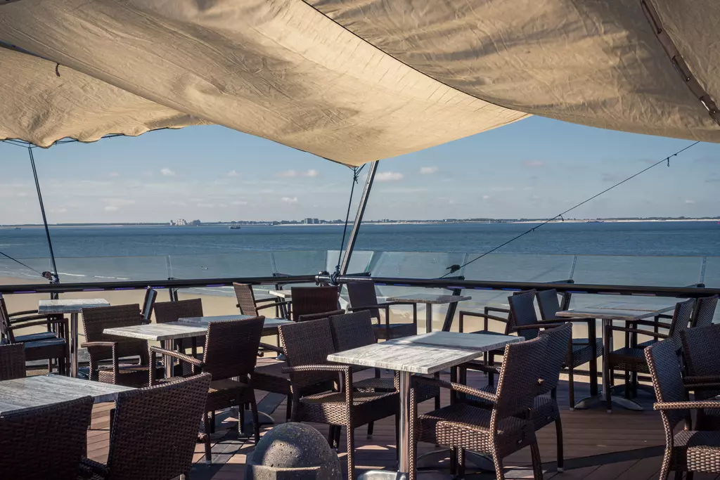 Tables and chairs on a terrace with a sunshade, overlooking the water and a clear sky in the background.