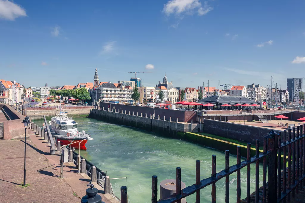 A harbor with a small boat, shallow water, and surrounding buildings. The sky is blue with a few clouds.