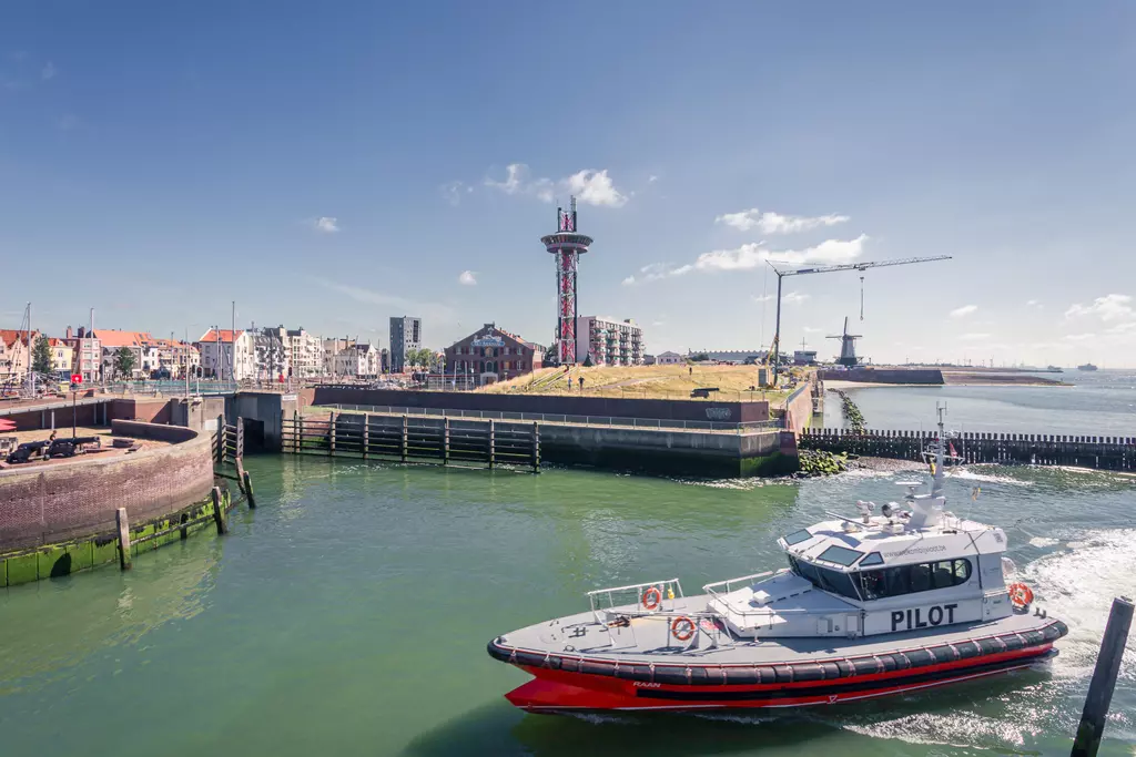 A pilot boat navigates through green water, with buildings and a lookout tower in the background.