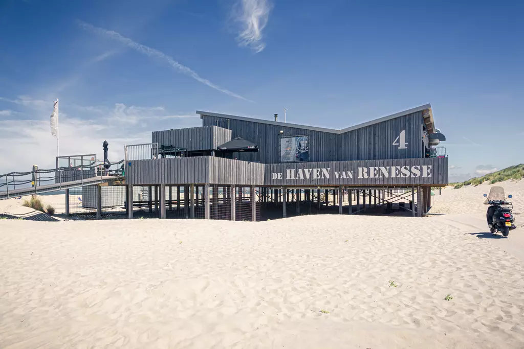 A modern wooden building on stilts with large windows, featuring the name De Haven van Renesse and a motorcycle parked nearby.