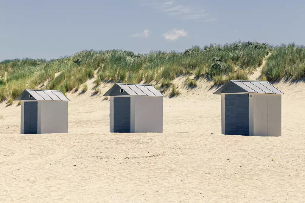 Three gray beach cabins stand on light sand, surrounded by gentle dunes and green grass in the background.